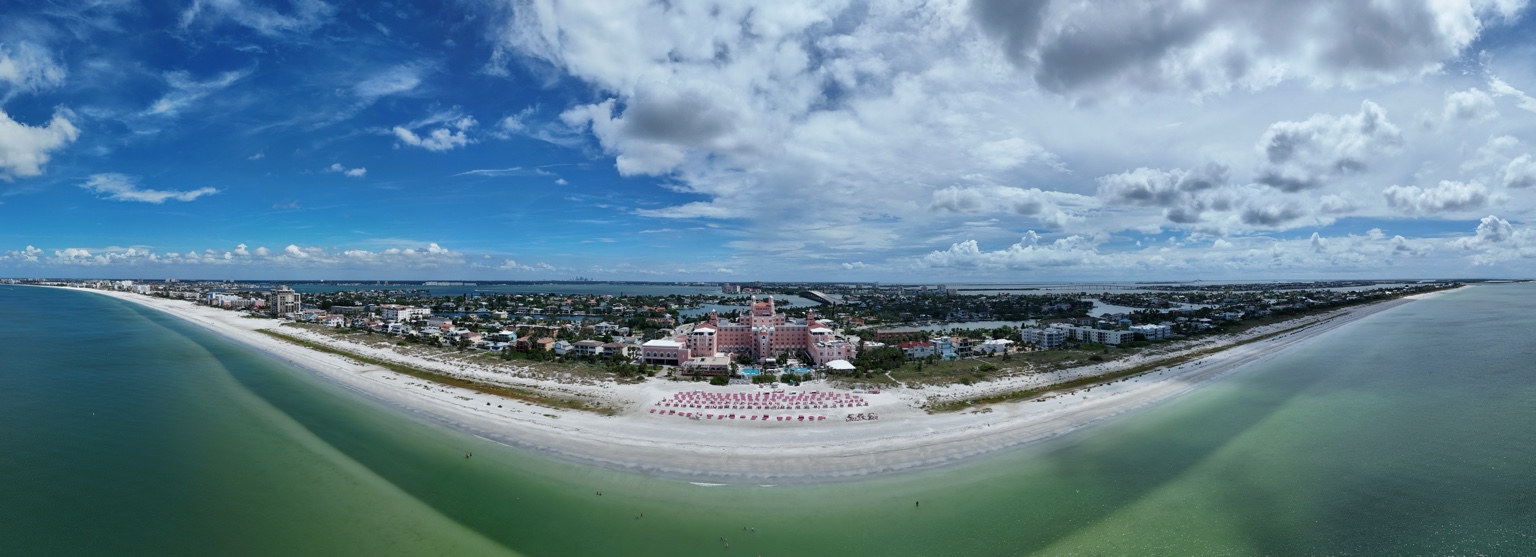 Pink beachfront resort centered along a sweeping white sand beach with green coastal waters and clouds overhead